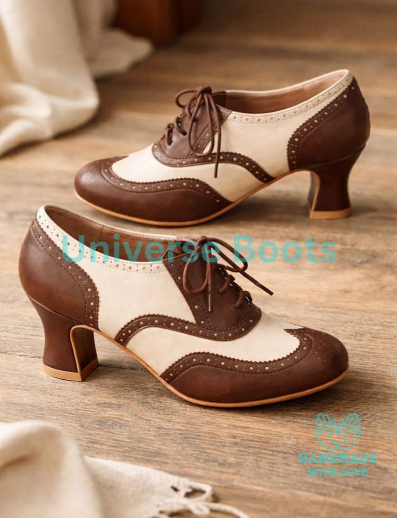 May include: A pair of two-tone brown and cream-colored oxford-style heels with a lace-up design. The shoes feature a rounded toe, a decorative perforated pattern, and a sturdy, stacked heel. The heels are approximately 2.5 inches high.