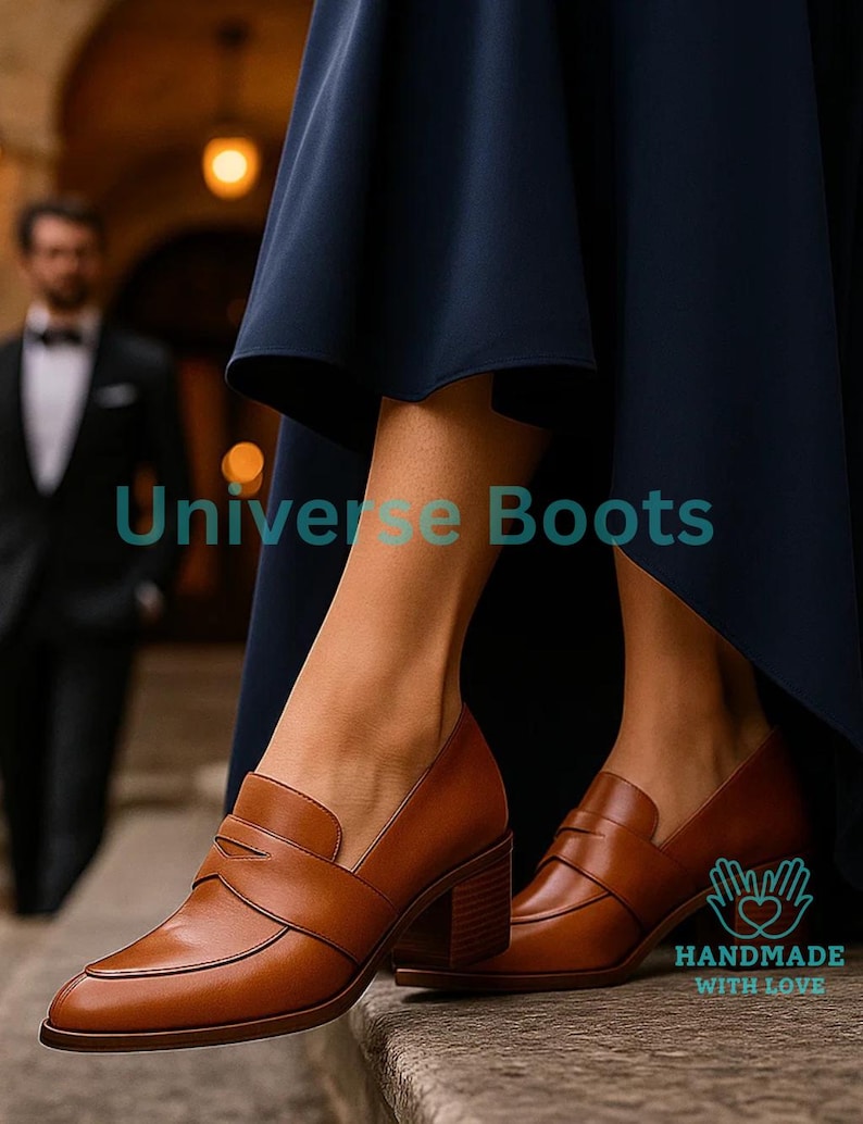 May include: Brown leather heeled loafers with a square toe and a strap across the top. The shoes have a block heel and are worn with a navy blue dress. The text "Universe Boots" is visible. The words "HANDMADE WITH LOVE" are also visible.