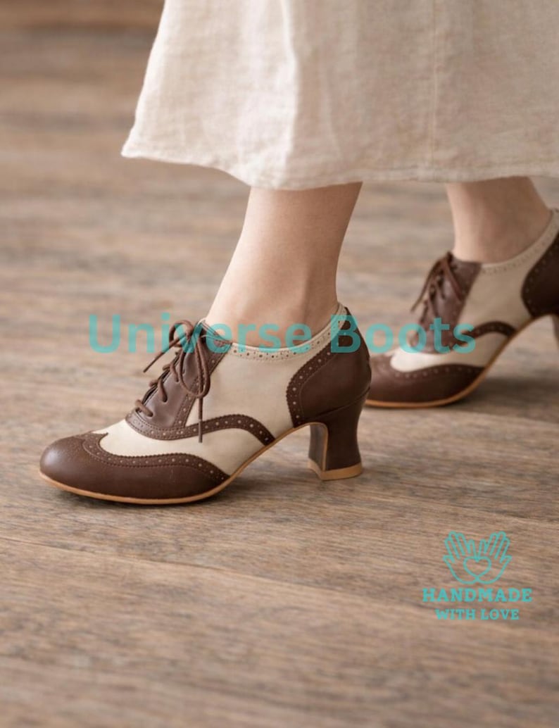 May include: Brown and cream-colored oxford-style shoes with a low heel. The shoes have a lace-up design and a classic wingtip pattern. The heels are approximately 2 inches high. The shoes are made of leather and have a vintage aesthetic.