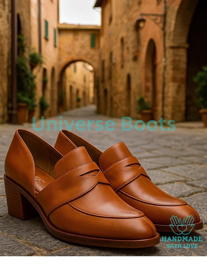 May include: A pair of brown leather heeled loafers. The shoes feature a classic design with a pointed toe and a block heel. The image includes the text "Universe Boots" and "HANDMADE WITH LOVE".