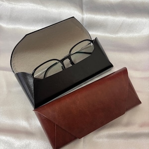 Può includere: Astuccio nero per occhiali aperto con occhiali dalla montatura nera all'interno. Sotto, un astuccio in pelle marrone chiuso. Gli astucci sono su una superficie bianca strutturata.