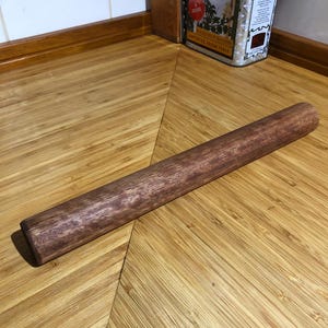 May include: A dark brown wooden rolling pin, cylindrical in shape, resting on a light brown bamboo surface. This kitchen utensil is used for flattening dough. The rolling pin is approximately 30 cm long.