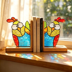 May include: A pair of stained glass bookends featuring colorful parrots with yellow, blue, and red textured glass panels. The parrots have large white eyes and are mounted on light-colored wooden bases, holding three books upright between them.