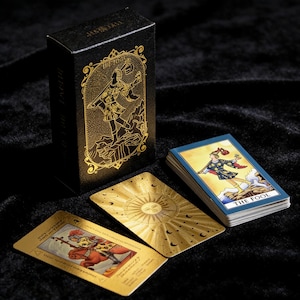 Op de afbeelding: Een zwarte tarotkaartdoos en -deck met gouden folie accenten. De doos toont een illustratie van een figuur en de kaarten tonen gedetailleerde kunstwerken. De kaarten liggen op een donker fluwelen oppervlak.