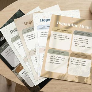 May include: A stack of "Dopamine Menu" printables in various colors, including black, beige, and white, are arranged on a wooden table. The menu features sections for "Sips & Sparks," "Tidy the Den," and "Sweet Treats." A pen and a mug are also on the table.
