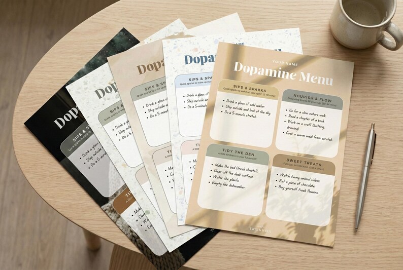 May include: A stack of "Dopamine Menu" printables in various colors, including black, beige, and white, are arranged on a wooden table. The menu features sections for "Sips & Sparks," "Tidy the Den," and "Sweet Treats." A pen and a mug are also on the table.