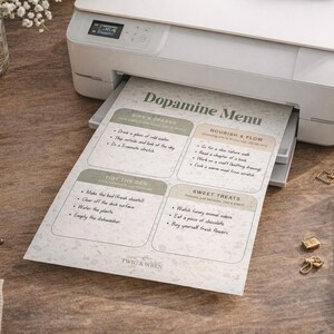 May include: A white printer printing a "Dopamine Menu" on paper. The menu features sections for "Birds & Sparks", "Nourish & Flow", "Tidy the Den", and "Sweet Treats". The scene includes a coffee cup, candle, and stationery.