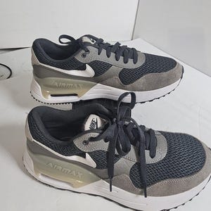May include: Pair of Nike Air Max sneakers. The shoes feature a black mesh upper with gray suede overlays and a white sole. The Nike swoosh is white, and the Air Max logo is visible on the side. The laces are black.