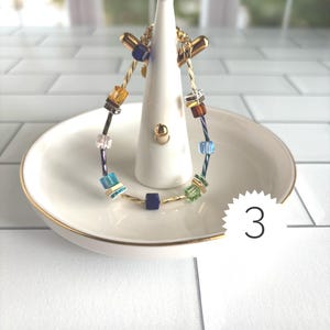 Puede incluir: Una pulsera de alambre trenzado en tono dorado con un surtido de cuentas cuadradas multicolores en tonos azul, verde, &aacute;mbar y transparente, acentuadas con peque&ntilde;os espaciadores met&aacute;licos. La pulsera se exhibe en un soporte de cer&aacute;mica blanca.