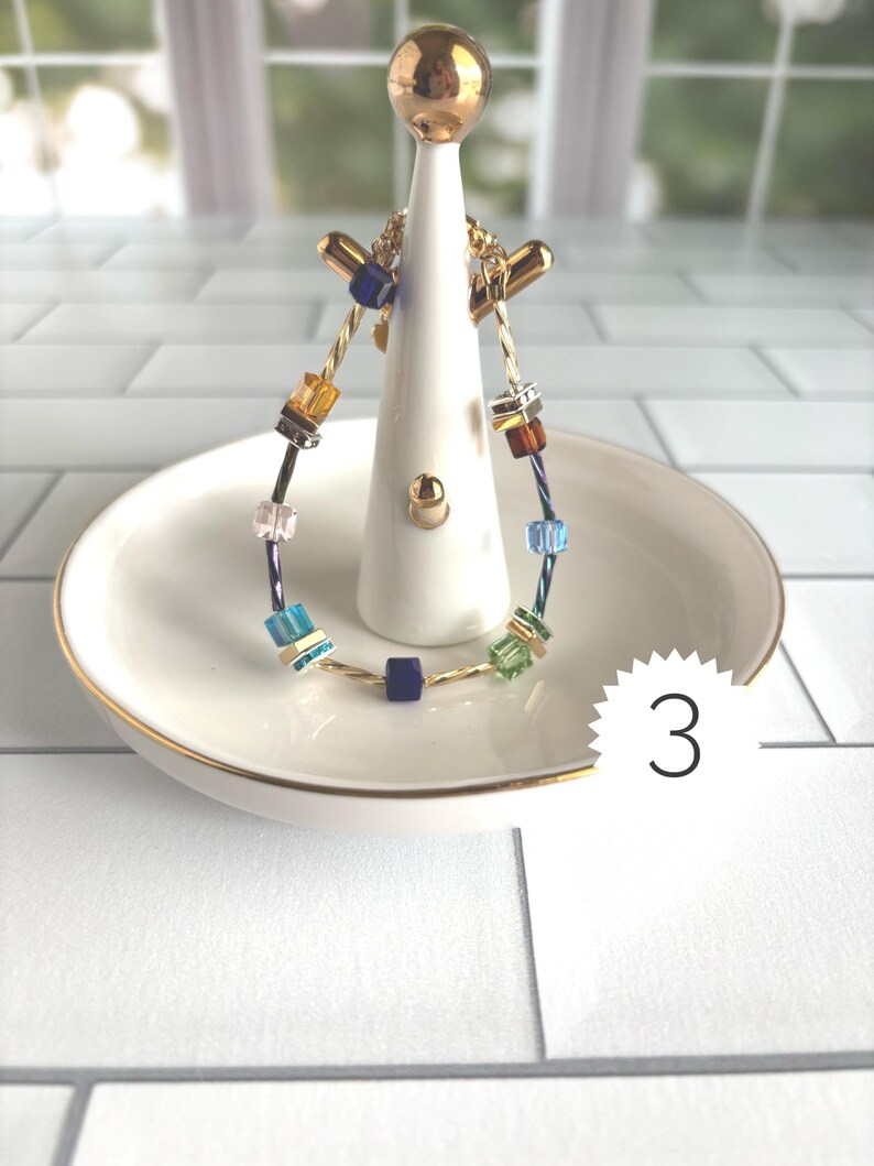 Puede incluir: Una pulsera de alambre trenzado en tono dorado con un surtido de cuentas cuadradas multicolores en tonos azul, verde, &aacute;mbar y transparente, acentuadas con peque&ntilde;os espaciadores met&aacute;licos. La pulsera se exhibe en un soporte de cer&aacute;mica blanca.