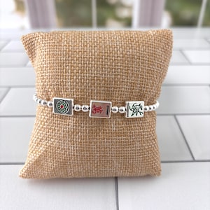 May include: A stretch bracelet featuring small silver-toned spherical beads interspersed with three square tiles. The tiles display colorful mahjong-style engravings, including a green floral pattern, a red character, and a green bamboo design.