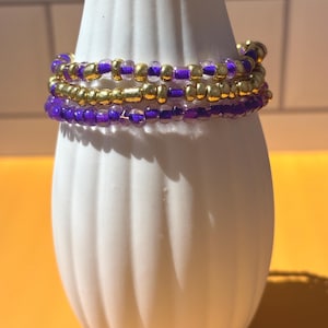 May include: Three beaded bracelets. The bracelets are wrapped around a white vase. The beads are a mix of gold and purple. The bracelets are stacked on top of each other. The background is a solid orange color.