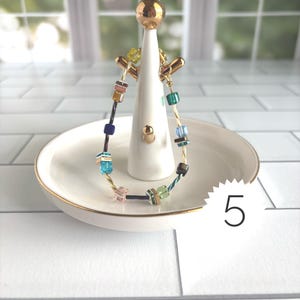 Puede incluir: Una pulsera delicada que presenta una mezcla de peque&ntilde;as cuentas cuadradas multicolores en tonos azul, verde, rosa y marr&oacute;n, ensartadas en un fino alambre dorado. La pulsera se exhibe en un soporte de cer&aacute;mica blanca para joyas con una parte superior dorada.
