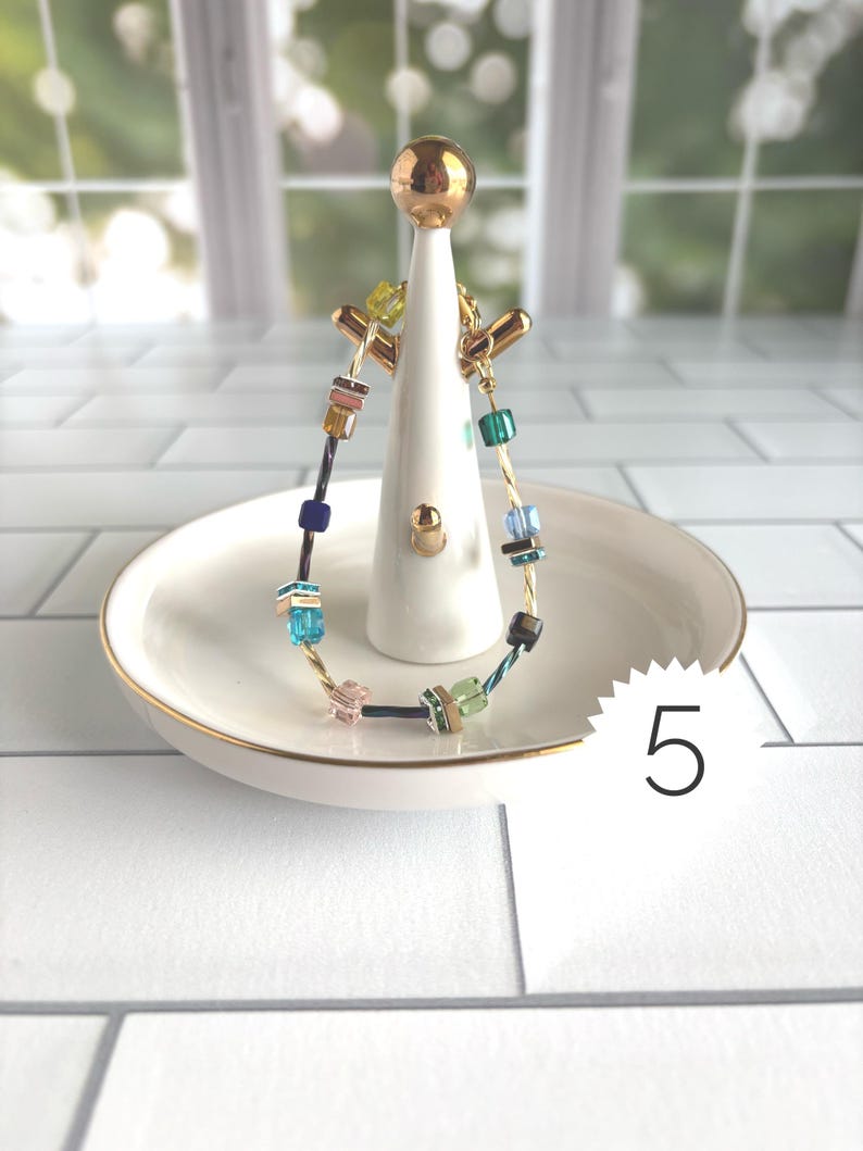 Puede incluir: Una pulsera delicada que presenta una mezcla de peque&ntilde;as cuentas cuadradas multicolores en tonos azul, verde, rosa y marr&oacute;n, ensartadas en un fino alambre dorado. La pulsera se exhibe en un soporte de cer&aacute;mica blanca para joyas con una parte superior dorada.