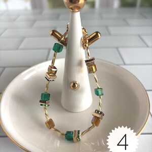 Puede incluir: Una pulsera de alambre trenzado en tono dorado con cuentas cuadradas alternas en tonos verde esmeralda, &aacute;mbar y plata met&aacute;lica. La pulsera se muestra en un soporte blanco e incluye una etiqueta circular blanca con el n&uacute;mero 4 impreso en negro.
