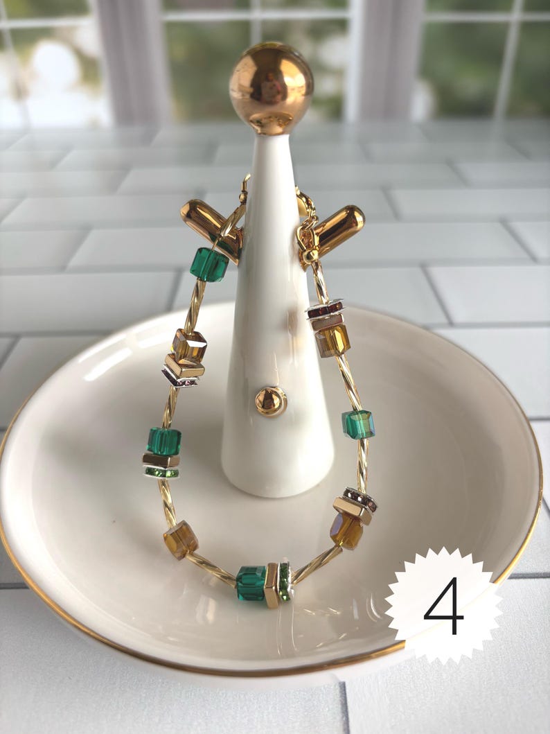 Puede incluir: Una pulsera de alambre trenzado en tono dorado con cuentas cuadradas alternas en tonos verde esmeralda, &aacute;mbar y plata met&aacute;lica. La pulsera se muestra en un soporte blanco e incluye una etiqueta circular blanca con el n&uacute;mero 4 impreso en negro.