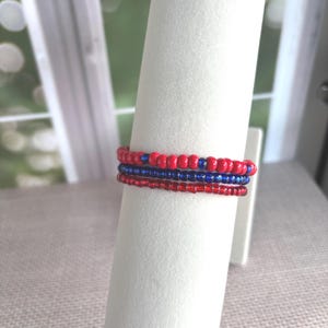 May include: A display of three beaded bracelets in red and blue. The bracelets are made of small, round beads and are stacked on a white cylindrical form. The background is a soft, neutral color, with a window visible in the distance.