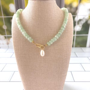 May include: A necklace featuring a strand of round, pale green beads. The necklace includes a gold-toned metal clasp positioned at the front, from which a single, white, teardrop-shaped pearl pendant hangs as a central focal point.