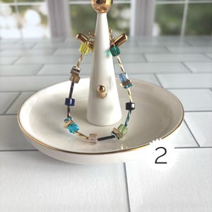 Puede incluir: Una pulsera con una banda de alambre dorado trenzado acentuada con cuentas cuadradas multicolores en tonos amarillo, verde, azul y rosa. La pulsera se exhibe en un soporte de cer&aacute;mica blanca para anillos con una parte superior dorada y una etiqueta n&uacute;mero 2.