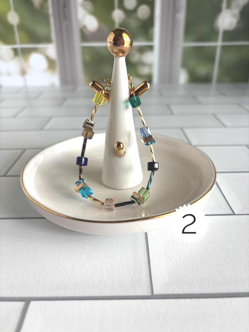 Puede incluir: Una pulsera con una banda de alambre dorado trenzado acentuada con cuentas cuadradas multicolores en tonos amarillo, verde, azul y rosa. La pulsera se exhibe en un soporte de cer&aacute;mica blanca para anillos con una parte superior dorada y una etiqueta n&uacute;mero 2.