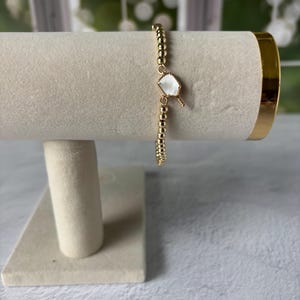 May include: A gold-toned beaded bracelet featuring a single, irregular-shaped white stone charm with a gold-toned metal border. The bracelet is composed of small, uniform metallic spheres strung together to form a continuous circular band.