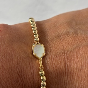 May include: Gold beaded bracelet featuring a pearlescent, shield-shaped charm. The charm is outlined in gold and accented with a small, sparkling stone. The bracelet is made of small, round gold beads.