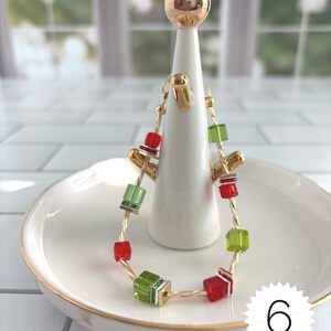 Puede incluir: Una pulsera de alambre trenzado en tono dorado con cuentas c&uacute;bicas alternas en rojo y verde lima. La pulsera se exhibe en un soporte c&oacute;nico blanco con una esfera dorada en la parte superior, sobre un plato blanco con borde dorado. El n&uacute;mero 6 es visible.