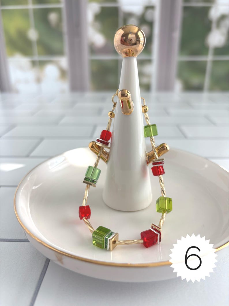 Puede incluir: Una pulsera de alambre trenzado en tono dorado con cuentas c&uacute;bicas alternas en rojo y verde lima. La pulsera se exhibe en un soporte c&oacute;nico blanco con una esfera dorada en la parte superior, sobre un plato blanco con borde dorado. El n&uacute;mero 6 es visible.
