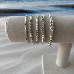 May include: A display of silver beaded bracelets. The bracelets vary in bead size, from small to larger, and are strung together. They are presented on a white, cylindrical display stand against a blurred ocean backdrop.