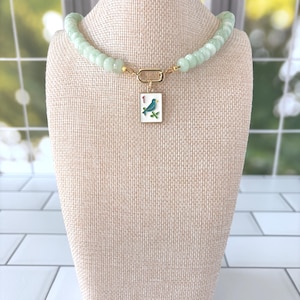 Puede incluir: Un collar con una hilera de cuentas cilíndricas acanaladas de color verde pálido y un cierre dorado. Un colgante rectangular blanco cuelga del centro, mostrando un gráfico de un pájaro azul, el número uno en rojo y hojas de bambú verdes.