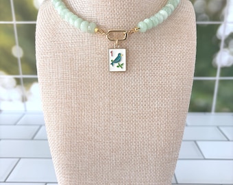Puede incluir: Un collar con una hilera de cuentas cilíndricas acanaladas de color verde pálido y un cierre dorado. Un colgante rectangular blanco cuelga del centro, mostrando un gráfico de un pájaro azul, el número uno en rojo y hojas de bambú verdes.