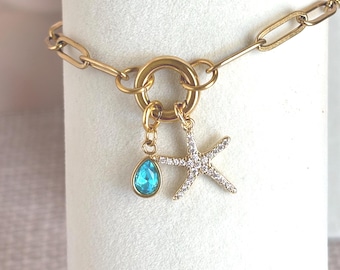 May include: A gold-toned chain bracelet featuring a circular clasp with two hanging charms: a teardrop-shaped pendant with a bright blue stone and a starfish-shaped pendant encrusted with small, clear, sparkling white crystals.