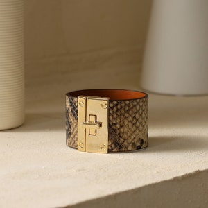 K&ouml;nnte beinhalten: Ein breites braunes Lederarmband mit einem goldfarbenen Verschluss und einem Schlangenhautmuster.