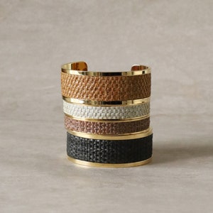 May include: Four stacked gold bracelets with woven brown, white, brown, and black bands.