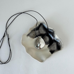 Puede incluir: Un collar con un colgante redondo de plata, de forma orgánica, que descansa sobre un plato de cerámica blanca con detalles negros.