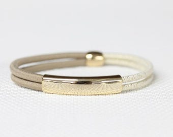 May include: A bracelet featuring two parallel cords, one in a light beige and the other in a cream color, connected by a polished gold-tone metal bar and a small matching gold-tone metal bead. The design is simple, sleek, and circular in shape.