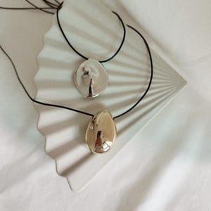 May include: Two necklaces with abstract, organic-shaped pendants. One pendant is silver and the other is gold. Both pendants are smooth and reflective. The necklaces are on a white background.