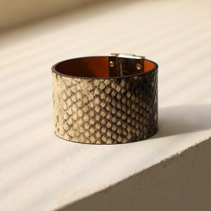 K&ouml;nnte beinhalten: Ein breites braunes Lederarmband mit Schlangenhautmuster und einem goldenen Verschluss.