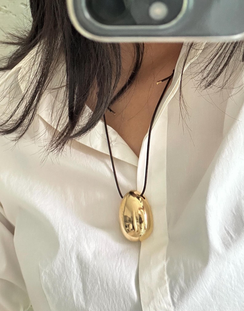 May include: A gold egg-shaped pendant hangs from a black cord necklace. The pendant is smooth and shiny.