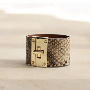 K&ouml;nnte beinhalten: Ein breites, goldfarbenes und braunes Lederarmband mit Schlangenhautmuster und einem goldenen Verschluss.