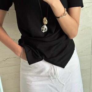 May include: A woman wearing a black t-shirt with a silver and gold pendant necklace and a white denim skirt.