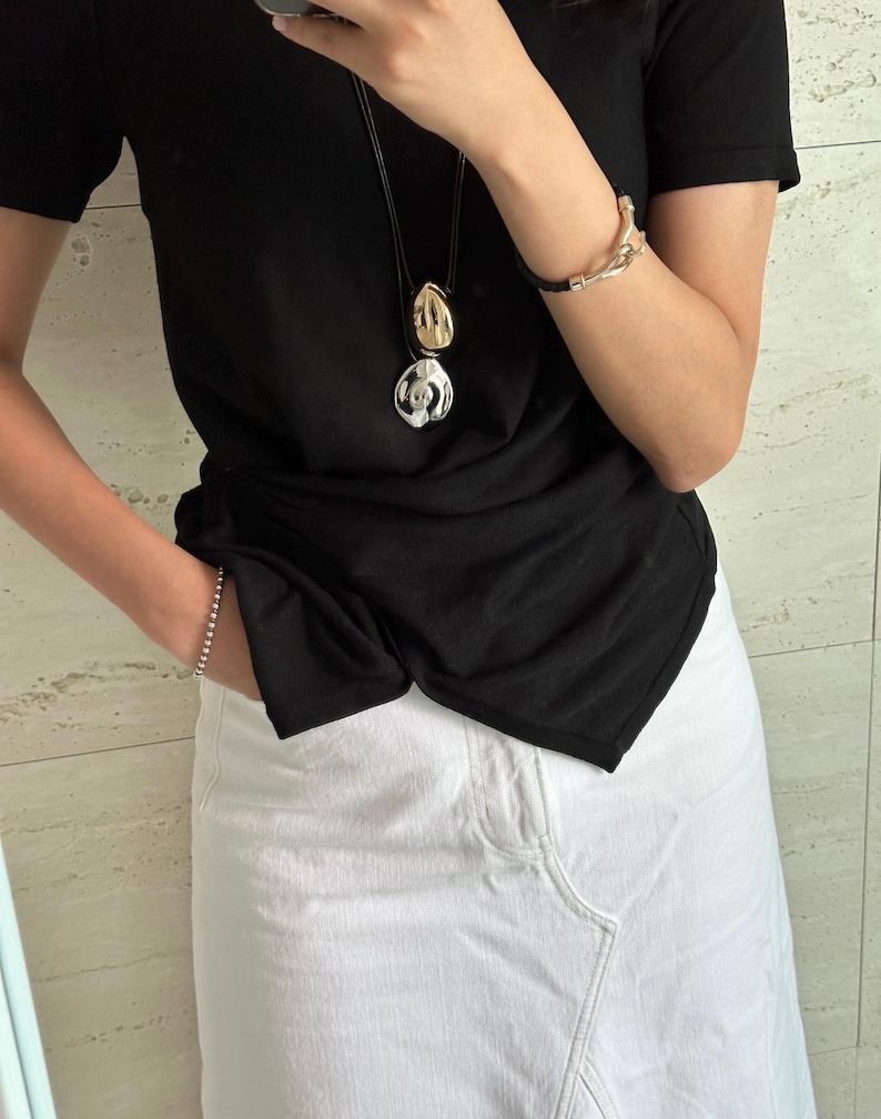 May include: A woman wearing a black t-shirt with a silver and gold pendant necklace and a white denim skirt.