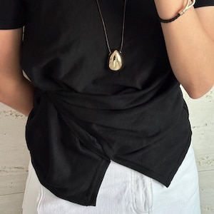 May include: A black t-shirt with a gold pendant necklace. The pendant is a teardrop shape. The shirt is tied at the waist and the person is wearing white denim shorts.