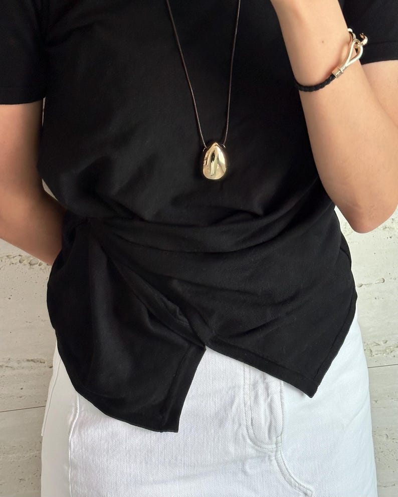 May include: A black t-shirt with a gold pendant necklace. The pendant is a teardrop shape. The shirt is tied at the waist and the person is wearing white denim shorts.