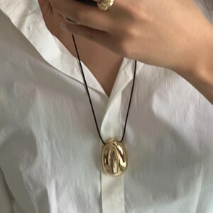 May include: A gold pendant necklace with a textured, abstract, round pendant. The necklace is on a black cord and is worn by a person wearing a white shirt.