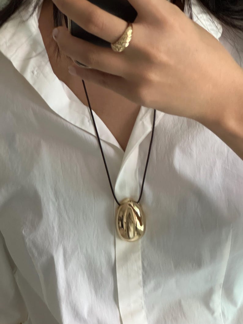 May include: A gold pendant necklace with a textured, abstract, round pendant. The necklace is on a black cord and is worn by a person wearing a white shirt.
