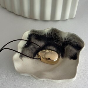 May include: A gold-coloured pendant necklace with a smooth, oval-shaped charm. The necklace is displayed on a white ceramic dish with black and white marbling.