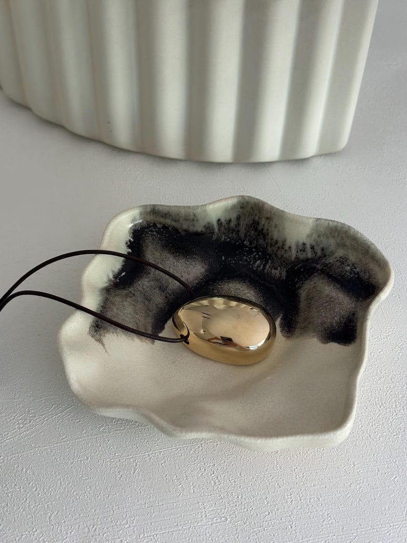 May include: A gold-coloured pendant necklace with a smooth, oval-shaped charm. The necklace is displayed on a white ceramic dish with black and white marbling.