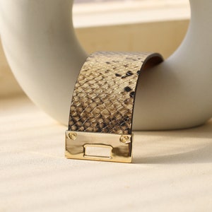 K&ouml;nnte beinhalten: Ein goldfarbenes Metallarmband mit einem braunen und schwarzen Schlangenhautmuster.