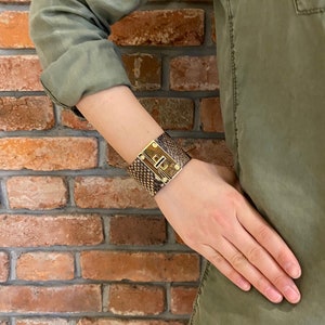 K&ouml;nnte beinhalten: Ein braunes Lederarmband mit einem goldenen Metallverschluss und einem Schlangenhautmuster.